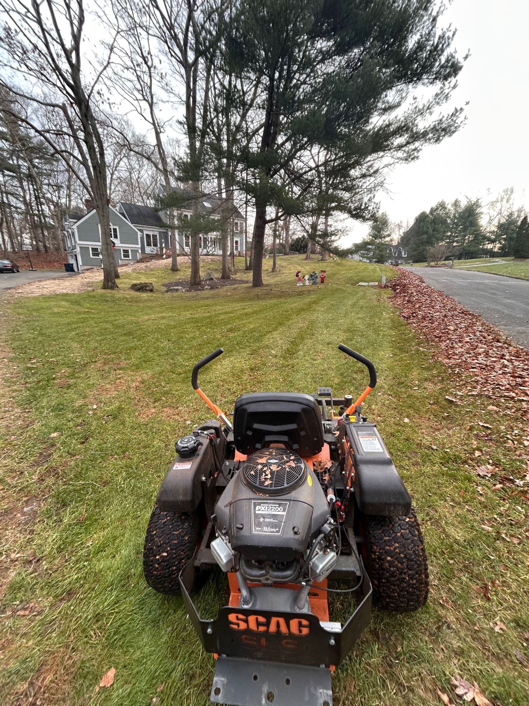 Zero-turn mower on a lawn beside a long row of leaves cleared to the driveway edge during a fall cleanup