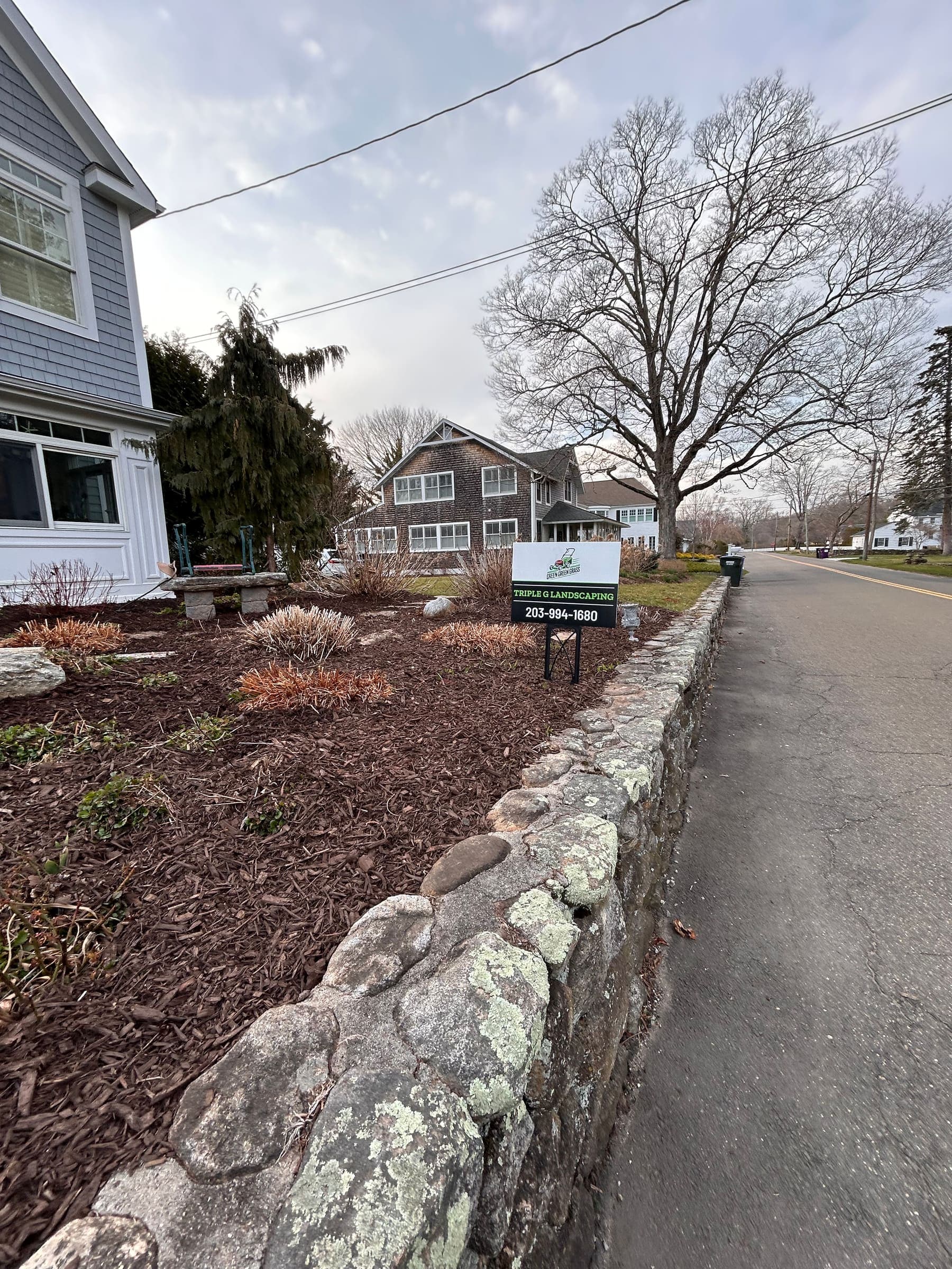 Mulched roadside bed behind a fieldstone wall with the Triple G Landscaping sign at the curb